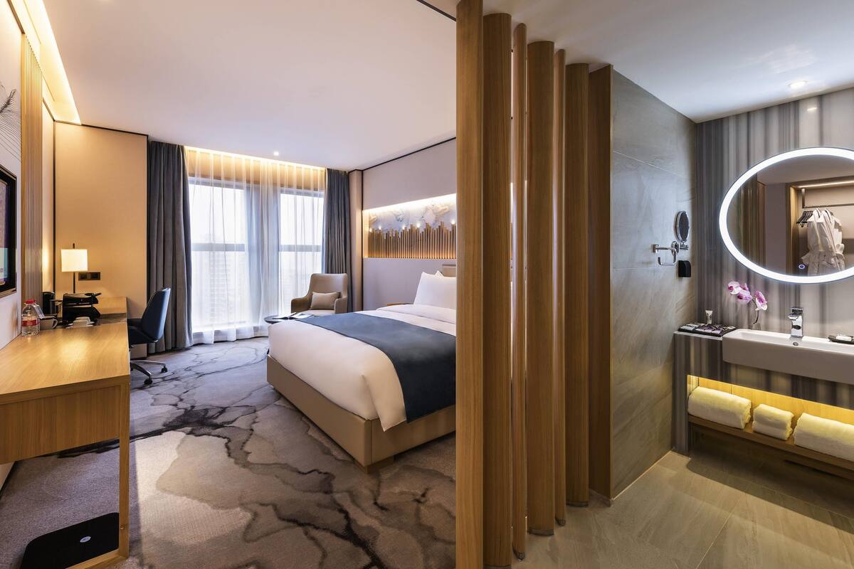 A modern hotel room with a bed, desk, chair, and a bathroom with a mirror and sink.