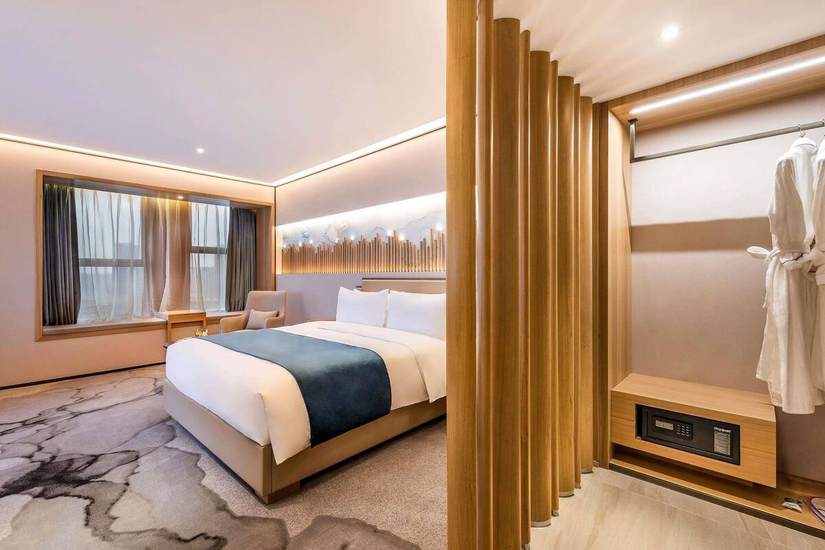 A hotel room with a bed, a seating area, a window with curtains, a wall-mounted light, and a robe rack.