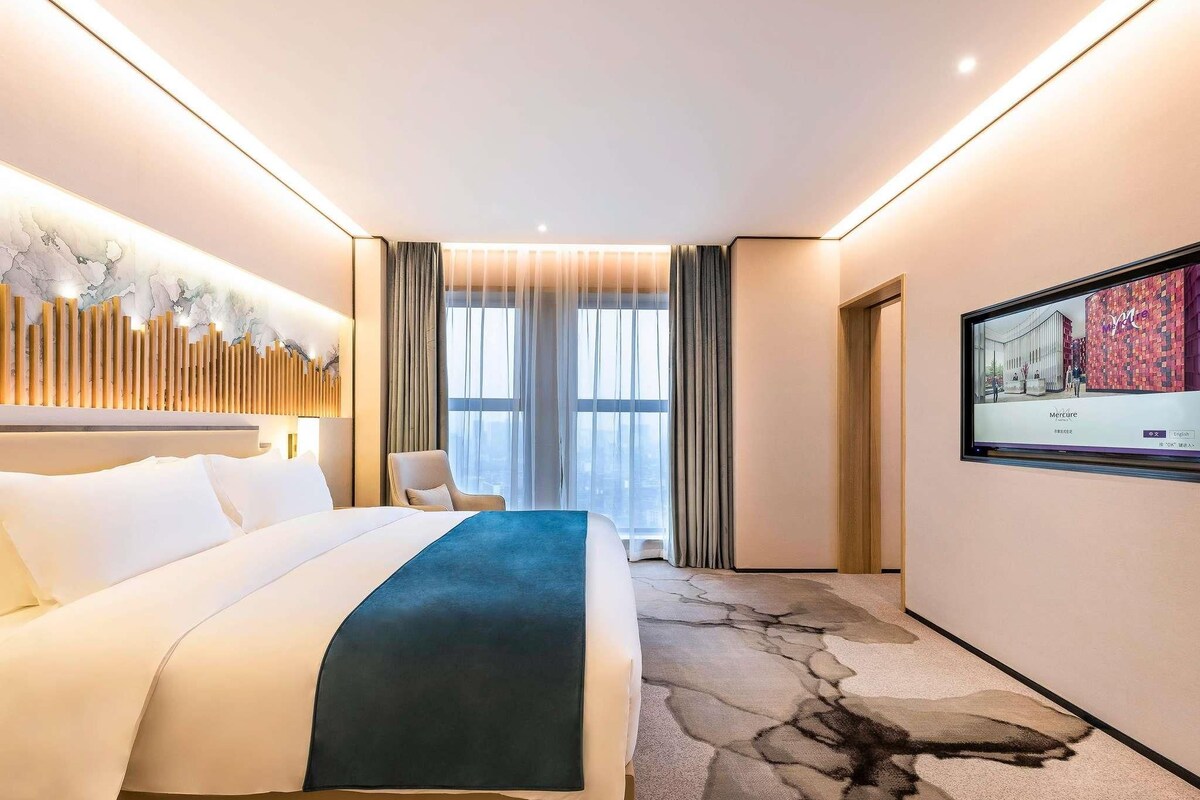 A hotel room with a bed, a chair, a TV, and a view of the city.