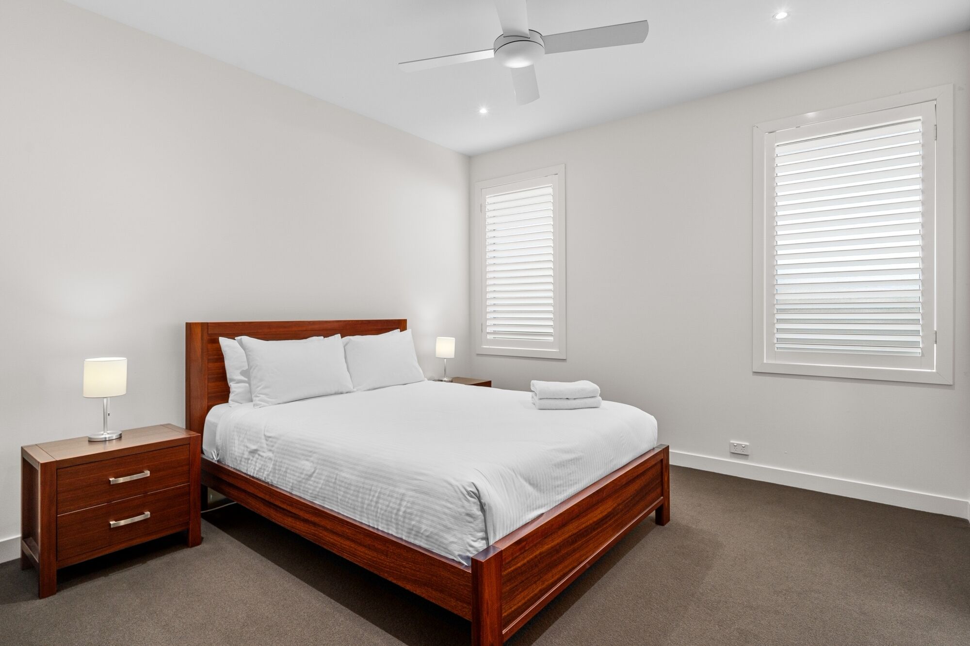 A bedroom with a wooden bed, white bedding, a nightstand with a lamp, and two windows with blinds.
