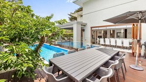 Et moderne udendørs poolområde med en træterrasse, et spisebord med stole og et overdækket loungeområde.