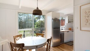 A modern kitchen with a round dining table, wooden chairs, a stainless steel refrigerator, and a view of the outdoors through large windows. - House One @ Potato Point (POTATO POINT)