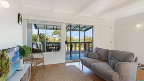 A modern living room with a large window, a sofa, a TV, and a wooden deck outside. - House One @ Potato Point (POTATO POINT)