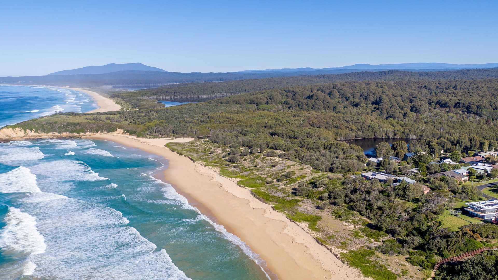 An aerial view of a coastal area with a sandy beach, dense forest, and a small cluster of buildings.