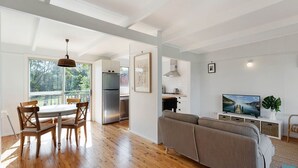 A modern kitchen with a built-in oven, a dining area with wooden chairs, a grey sofa, and a television on a stand. - House One @ Potato Point (POTATO POINT)