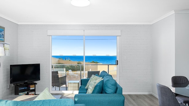 Ein Wohnzimmer mit großem Fenster mit Blick auf einen Strand, einem Flachbildfernseher und einer Sitzecke mit Sofa und Sesseln.