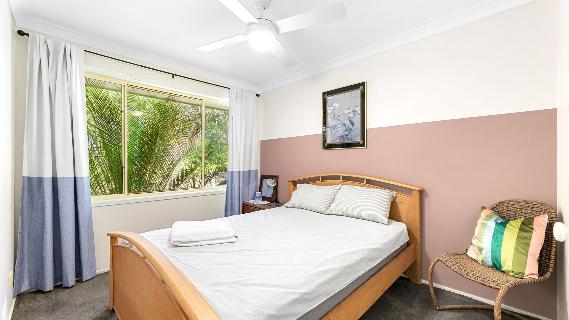 Una camera da letto con un letto, un comodino, una sedia, una ventola a soffitto e una finestra con tende.