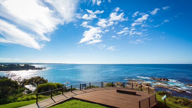 Une terrasse en bois, dotée d’une balustrade, qui offre une vue sur une zone côtière avec des rivages rocheux et un ciel bleu dégagé.
