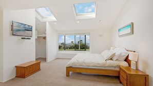 Une chambre à coucher comprenant un lit, une table de chevet, une commode, une fenêtre donnant sur l’extérieur et une lucarne au-dessus du lit.