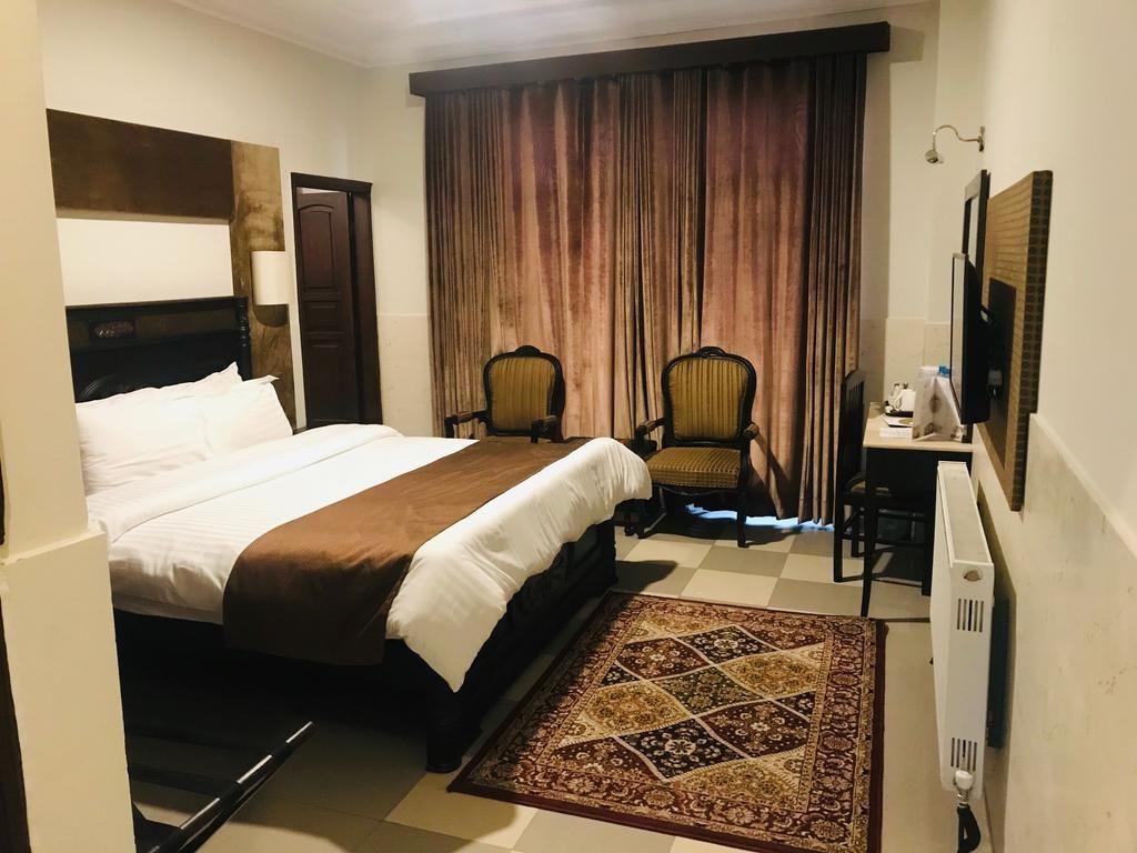 A hotel room with a large bed, two chairs, a desk, a television, and a patterned rug.