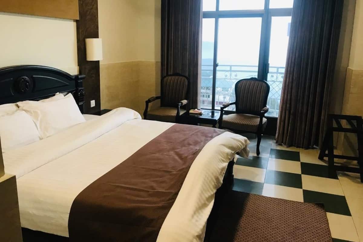 A hotel room with a large bed, two chairs, a view of the city through large windows, and a checkered floor.