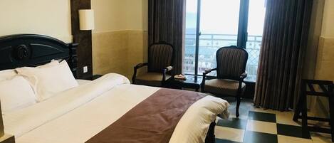 Habitación de hotel con una cama grande, dos sillones, vista de la ciudad a través de amplios ventanales y suelo a cuadros.