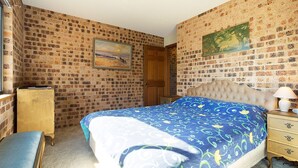 Een slaapkamer met een muur in baksteenmotief, een bed met een blauw bloemenbedekking, een houten nachtkastje met een lamp en een houten bankje.