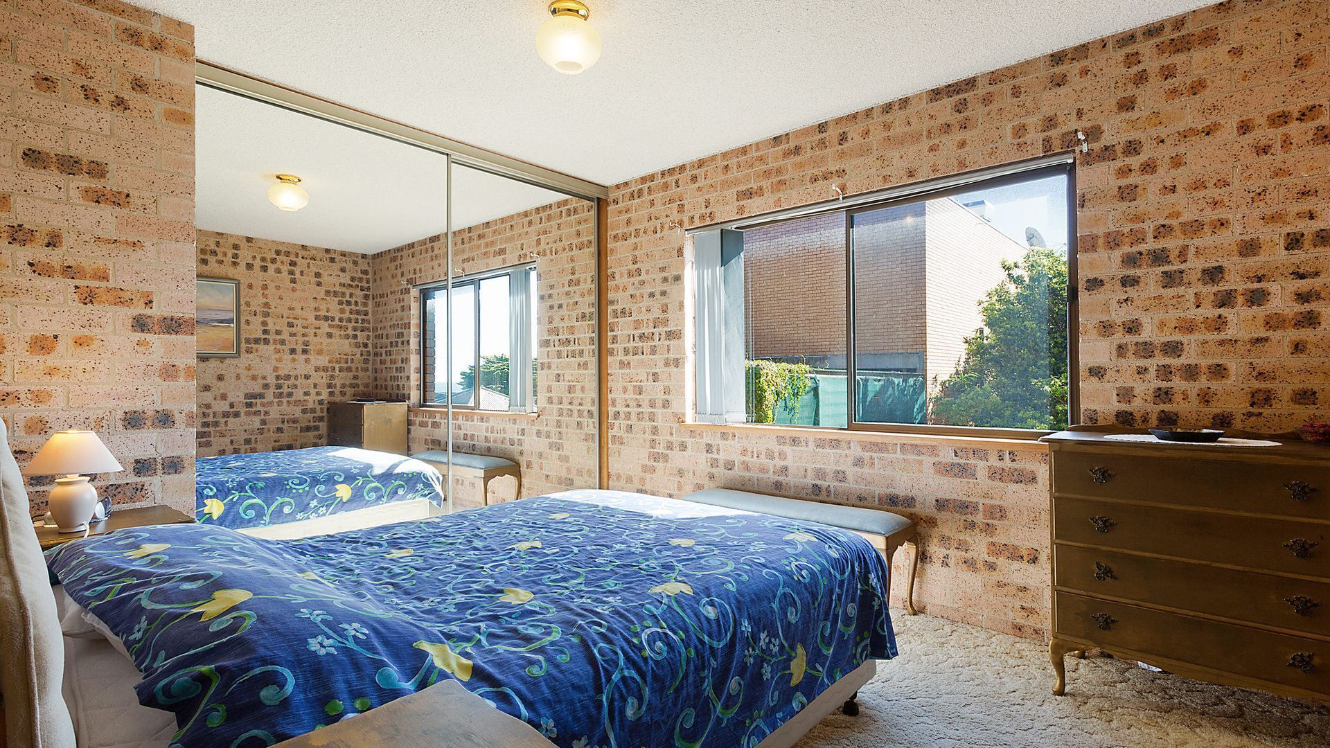 A bedroom with brick walls, a large mirror, a bed with a blue floral bedspread, a dresser, and a window with a view of greenery.