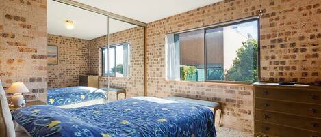 A bedroom with brick walls, a large mirror, a bed with a blue floral bedspread, a dresser, and a window with a view of greenery.