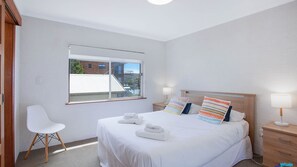 A hotel room with a large bed, a window with a view of buildings, a white chair, and a bedside table with a lamp.