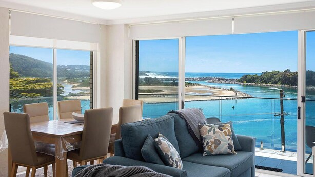 A spacious room with a large window offering a view of a coastal area, a dining table with chairs, and a sofa with decorative pillows.