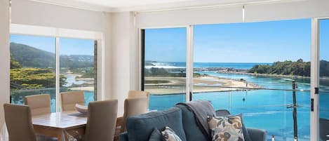 A spacious room with a large window offering a view of a coastal area, a dining table with chairs, and a sofa with decorative pillows.