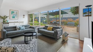 A modern living room with two grey sofas, a coffee table, and a patterned ottoman. Large windows offer a view of a wooden deck and garden.