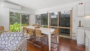 A modern kitchen with white cabinets, a dining area with a white table and wicker chairs, and large windows offering views of greenery.