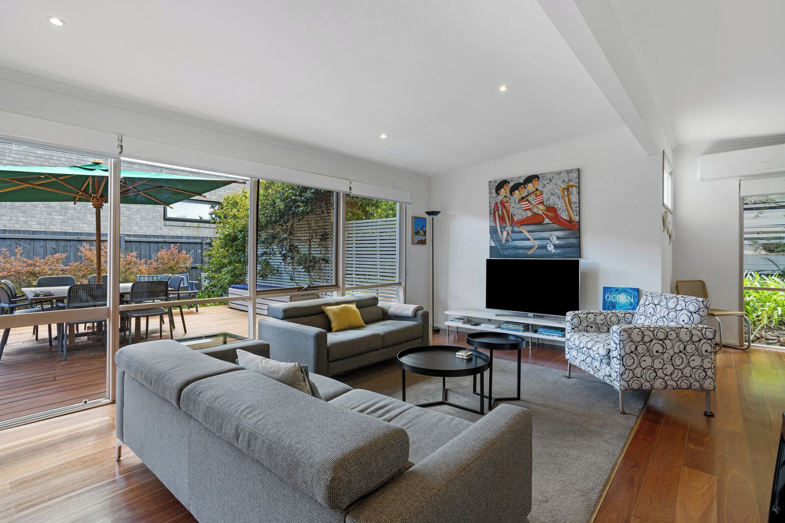 A modern living room with a grey sofa, a coffee table, and a TV. There is a balcony with outdoor furniture and a painting on the wall.