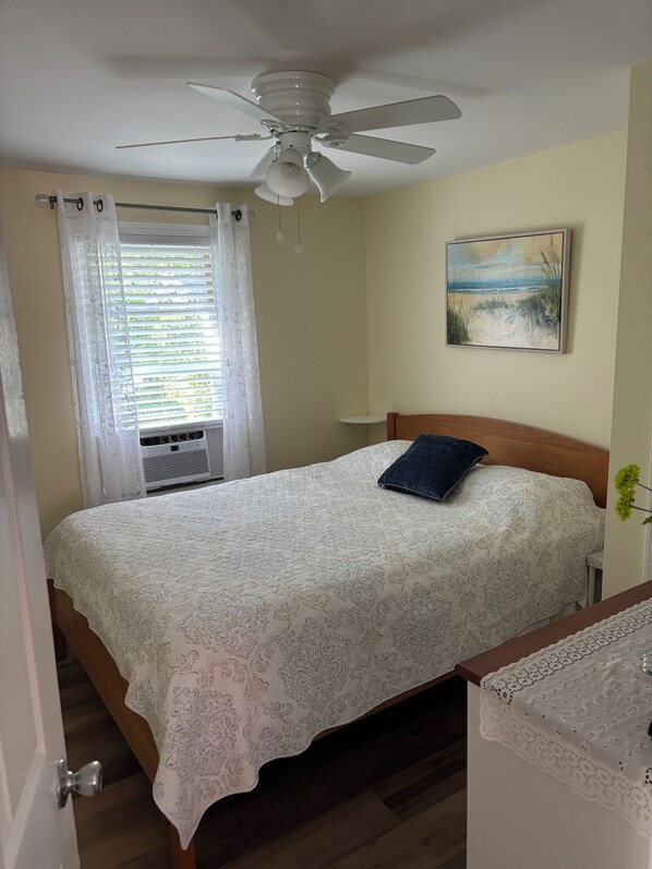 Un dormitorio con cama, ventilador de techo, ventana con cortinas, un cuadro en la pared y una mesita de noche con lámpara.