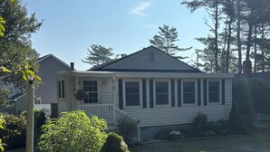 Ein einstöckiges Haus mit Veranda, ein kleiner Garten mit Topfpflanze und Steingrenze, und einem strahlend blauen Himmel.