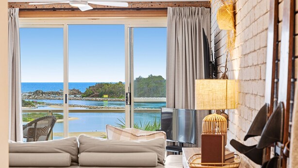 A room with a large sliding glass door leading to a beach view, a sofa, a TV, and a lamp. - Grand Pacific 1 Unit 1 - Ground Floor (NAROOMA)