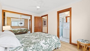 A bedroom with a bed, a bedside table, a lamp, a large mirror, and a door leading to a bathroom. - Grand Pacific 1 Unit 1 - Ground Floor (NAROOMA)