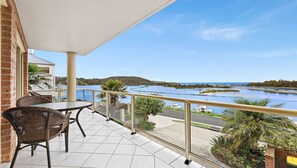 A balcony with a table and chairs, overlooking a river and distant hills. - Grand Pacific 1 Unit 1 - Ground Floor (NAROOMA)