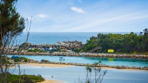 A coastal area with a rocky breakwater, a sandy beach, and a boat in the water. - Grand Pacific 2 Unit 1 - Ground Floor (NAROOMA)