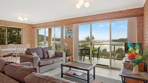 A spacious living area with a large sliding glass door leading to a balcony, a grey sofa, a coffee table, and a dining table with chairs. - Grand Pacific 2 Unit 1 - Ground Floor (NAROOMA)