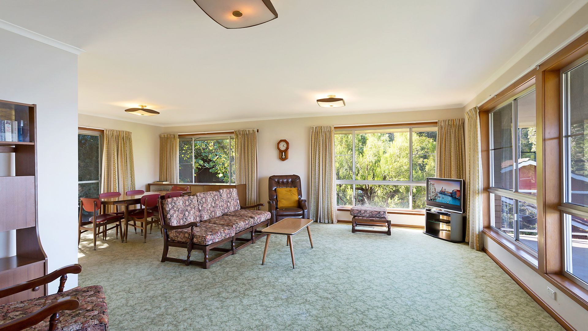 A spacious living room with a sofa, armchair, and a small table. There is a dining area with a table and chairs, and a television on a stand. Large windows provide natural light and views of greenery outside.