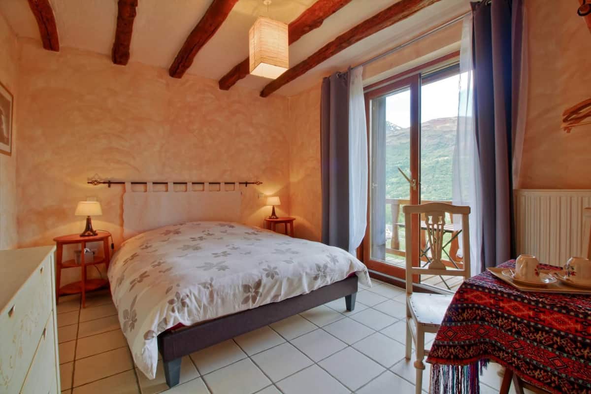 Un dormitorio con cama, dos mesitas de noche, una ventana con cortinas, un balcón con mesa y sillas, y vistas al exterior.