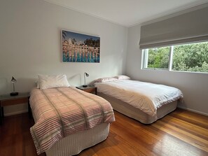 A bedroom with two beds, a nightstand with a lamp, a painting on the wall, and a window with a view of greenery. - BASS | 13 GUEST | TENNIS COURT | VIEWS | PORTSEA GOLF CLUB | LOCATION (Portsea)