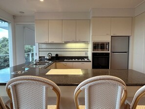 A modern kitchen with a central island, stainless steel appliances, and wicker chairs. - BASS | 13 GUEST | TENNIS COURT | VIEWS | PORTSEA GOLF CLUB | LOCATION (Portsea)