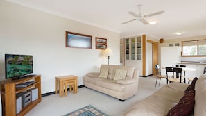 A living room with a beige sofa, a TV on a wooden stand, a small wooden table, a floor lamp, and a ceiling fan.