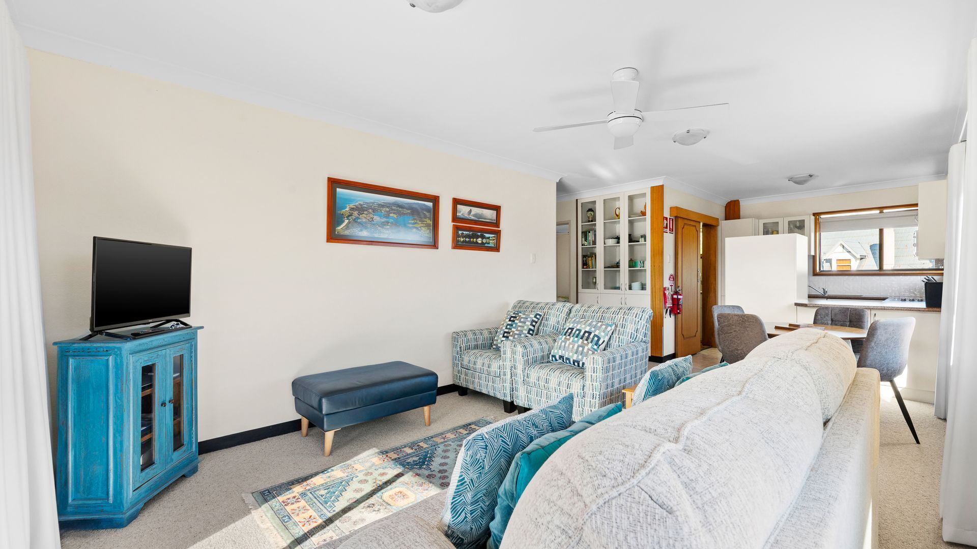 A living room with a television, sofa, and armchair, a kitchen area with a window, and a ceiling fan.