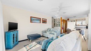 A living room with a television, sofa, and armchair, a kitchen area with a window, and a ceiling fan.
