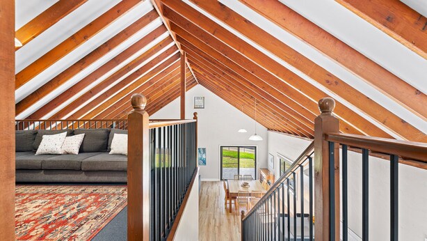 A loft-style room with exposed wooden beams, a gray sofa, and a dining area with a table and chairs. - Bush Retreat With Private Pool (NAROOMA)