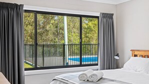 A hotel room with a large window, a bed with white linens, a wooden headboard, and a view of a swimming pool and greenery. - Bush Retreat With Private Pool (NAROOMA)