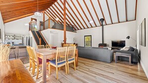 A spacious living area with a wooden dining table, chairs, a sofa, and a TV. A staircase leads to an upper level. - Bush Retreat With Private Pool (NAROOMA)