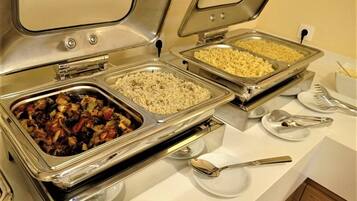 A buffet setup with various food items in metal containers, including pasta, rice, and a mixed vegetable dish.
