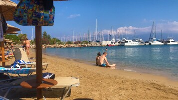 Una playa con sillas de descanso y sombrillas, un litoral rocoso y barcos en el agua.
