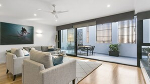 Un salon moderne comprenant un ensemble de canapés, un coin repas avec une table et des chaises, un ventilateur de plafond et de grandes fenêtres équipées de stores verticaux.