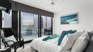 Une chambre dotée d’un grand lit, offrant une vue sur l’océan par la fenêtre, d’un ventilateur de plafond et d’un coin repas avec une table et des chaises.