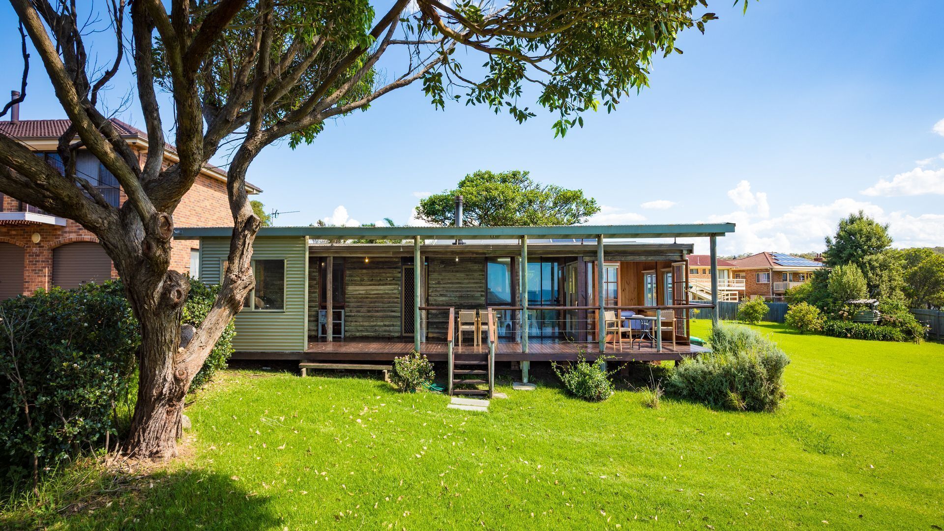 A modern log cabin with a deck, surrounded by a well-maintained lawn and a large tree.