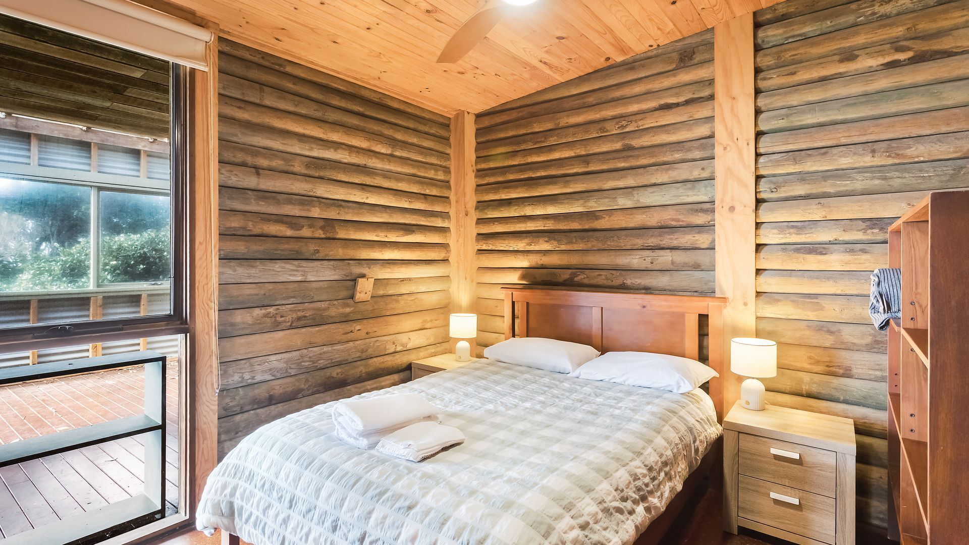 Un dormitorio tipo cabaña de madera con una cama, dos mesitas de noche con lámparas, un armario de madera y una ventana con vistas.