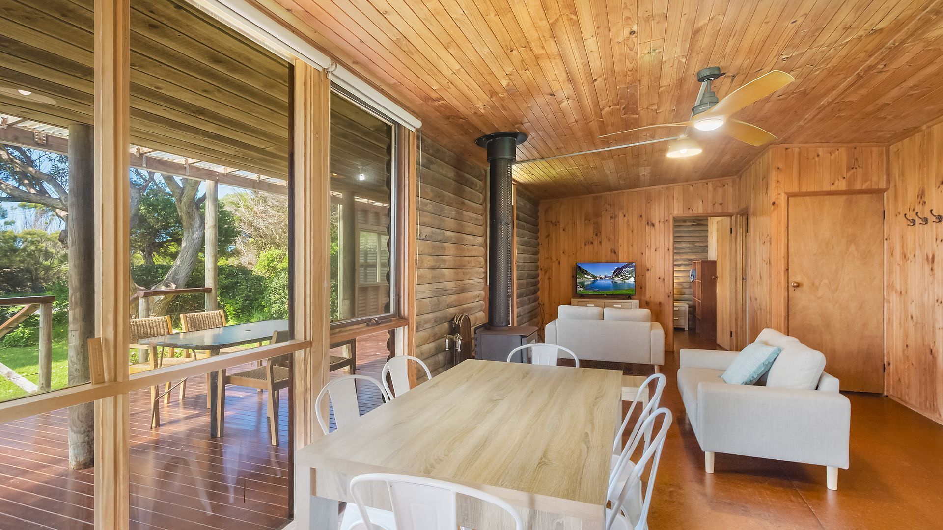 A wooden cabin interior with a dining table, white chairs, a sofa, and a TV.