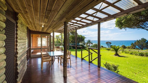 A wooden deck with a table and chairs, overlooking a grassy area and the ocean.